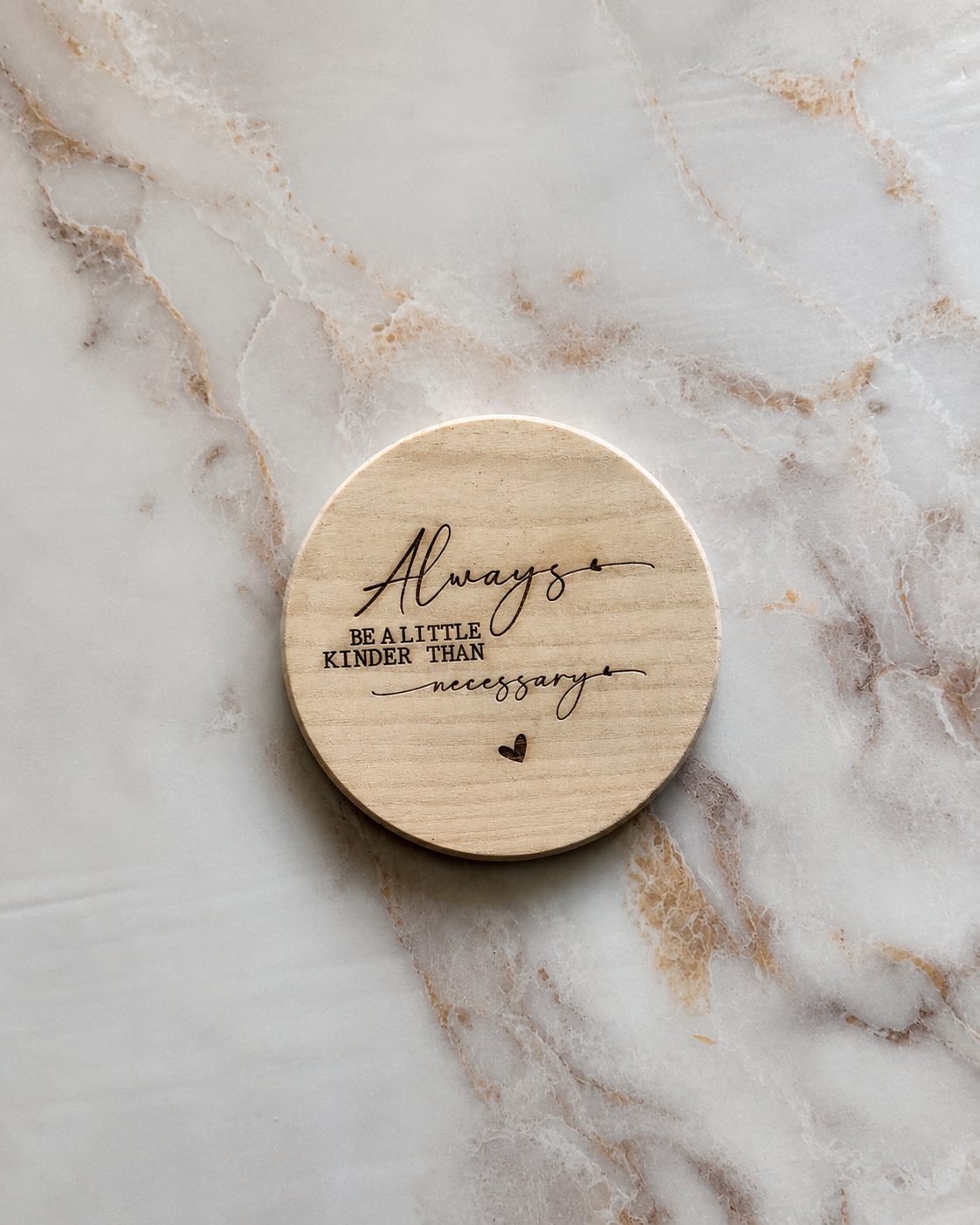 A round natural wood coaster with the engraved text 'Always be a little kinder than necessary' and a small heart, displayed on a white and beige marble surface.