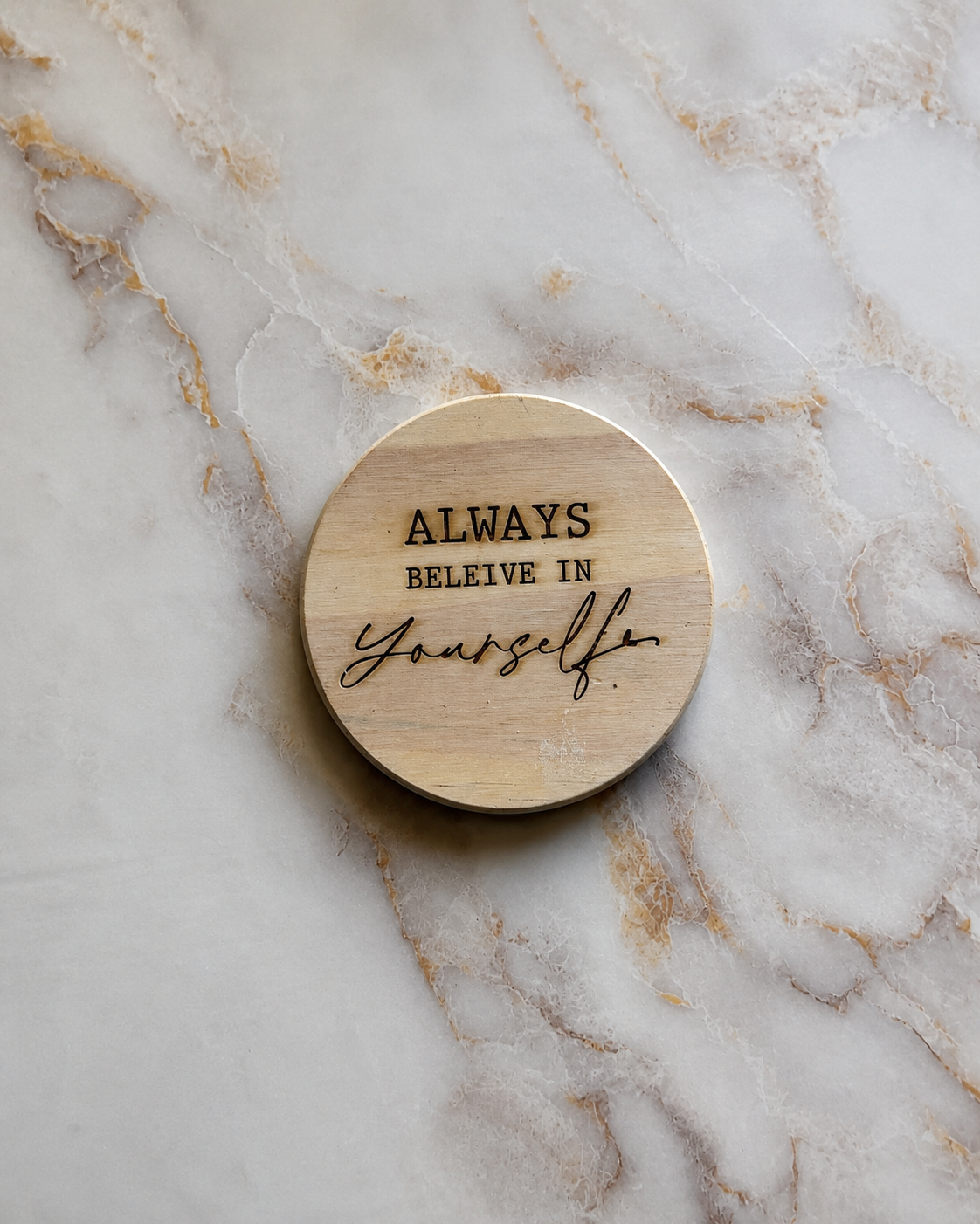 A round natural wood coaster with the engraved text 'ALWAYS BELEIVE IN Yourself.' resting on a white and gold marble surface.