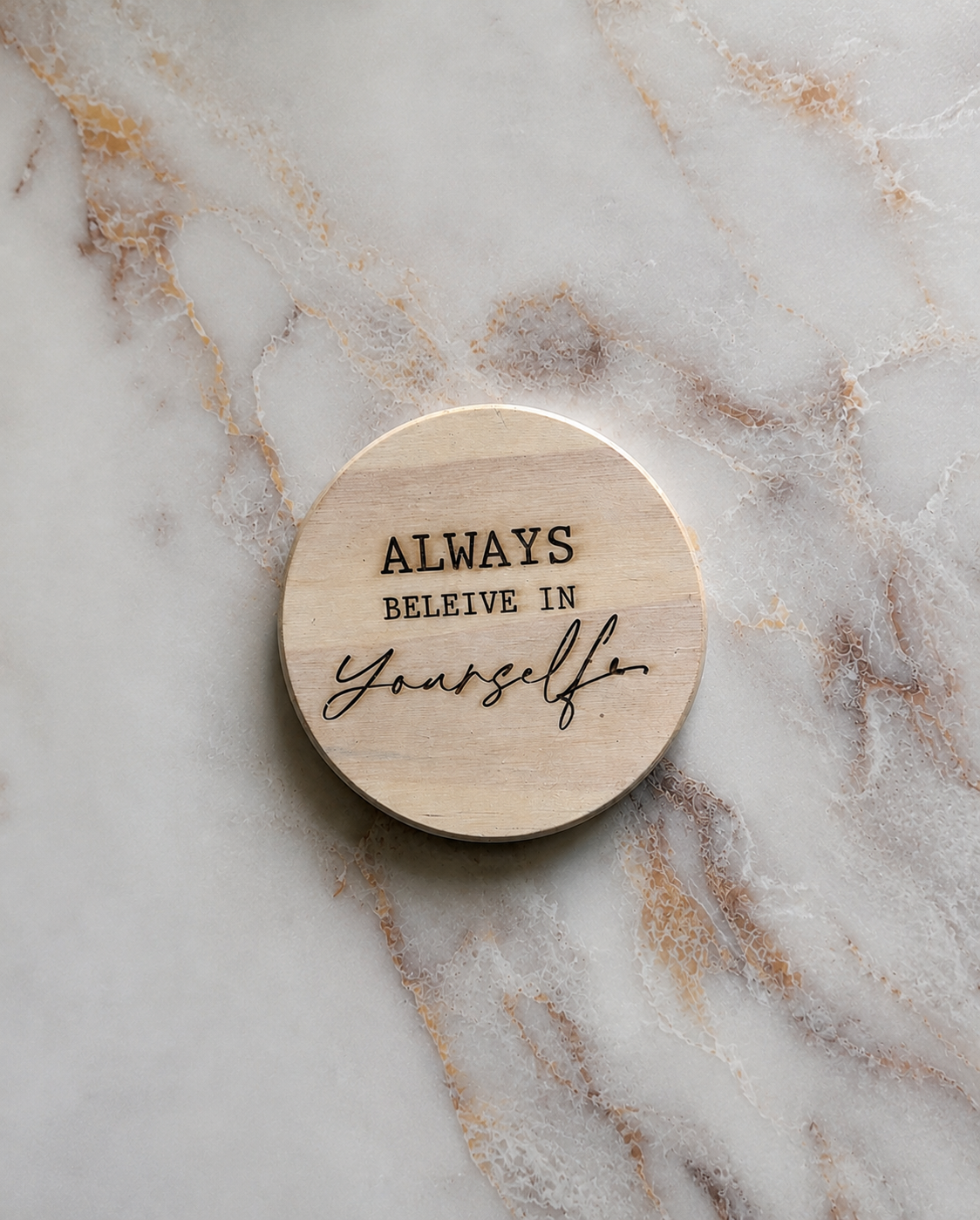 A round light wood coaster engraved with the words 'ALWAYS BELEIVE IN Yourself' in black lettering, placed on a white and beige marble surface.