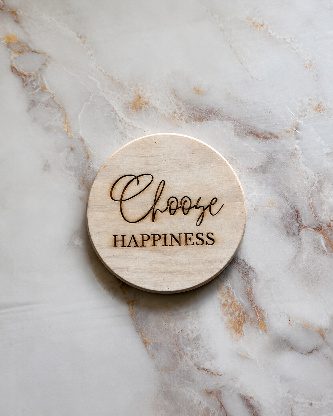 A round light wood coaster engraved with the words 'Choose HAPPINESS' in dark brown lettering, placed on a white and gold marble surface.
