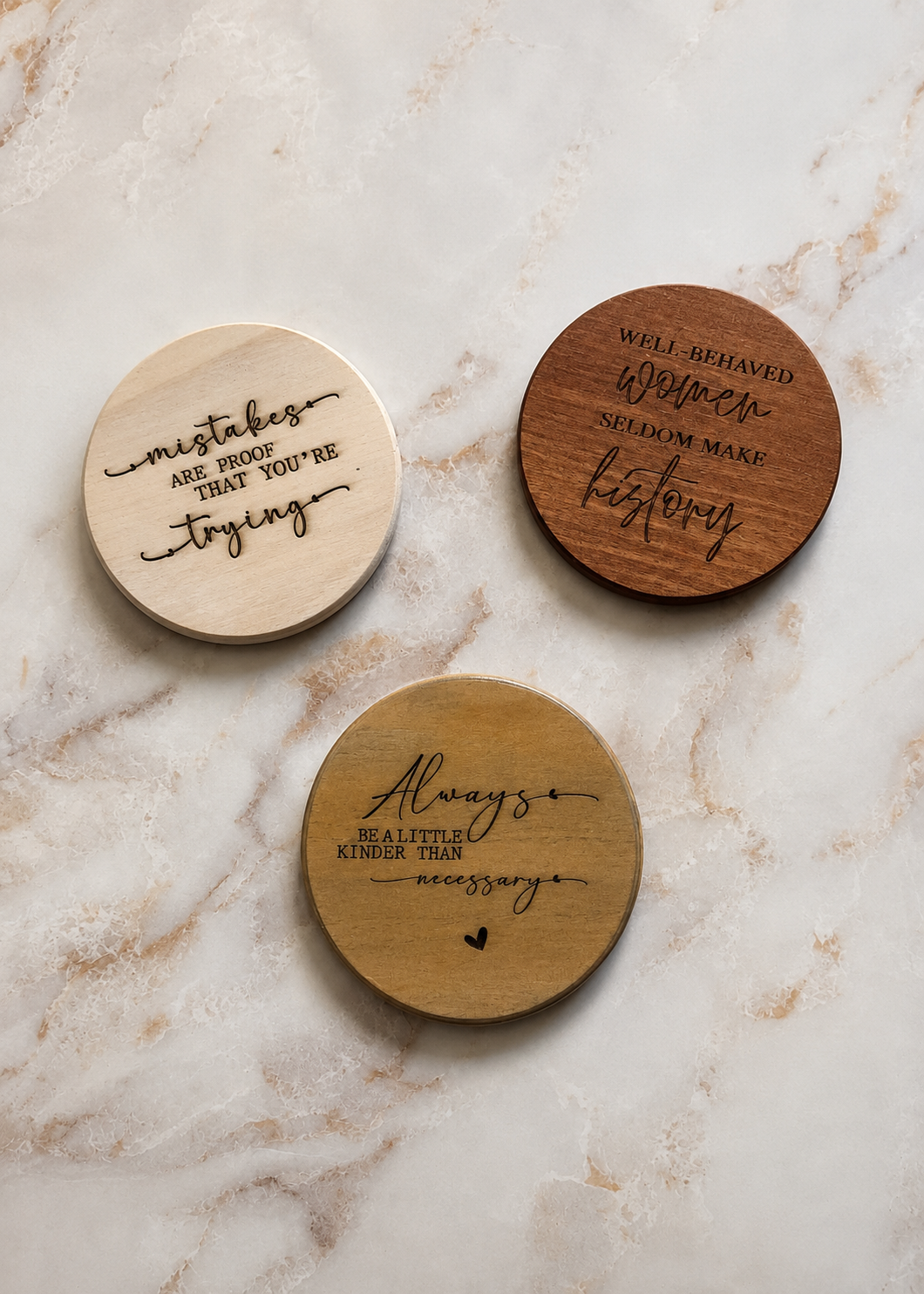 Three round wooden coasters with engraved inspirational quotes, displayed on a light beige marble surface. Coaster finshes are whitewashed, natural and traditional