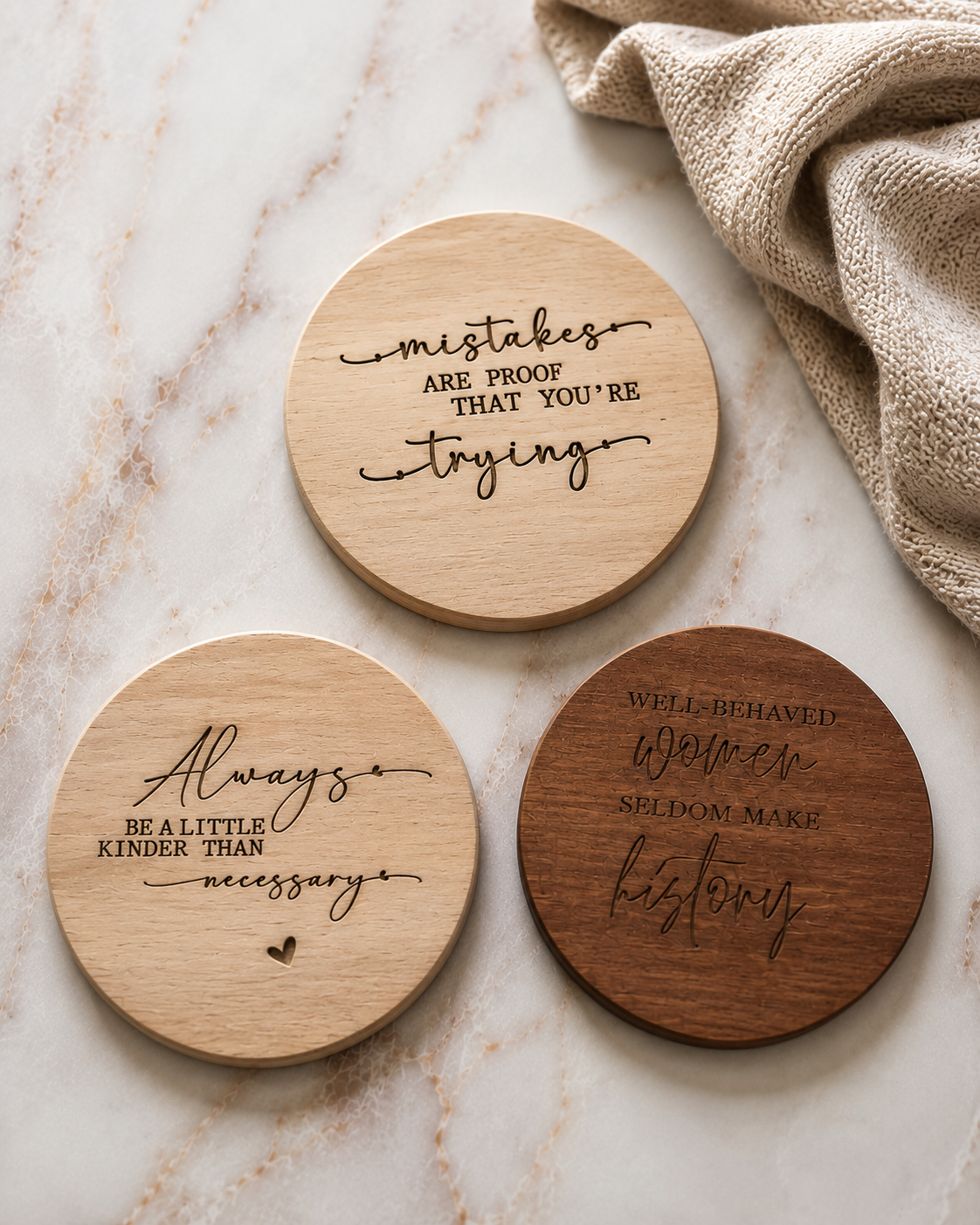 Three round wooden coasters with engraved inspirational quotes, arranged on a light marble surface next to a beige textured cloth. Two coasters are light natural wood and one is a darker brown wood.