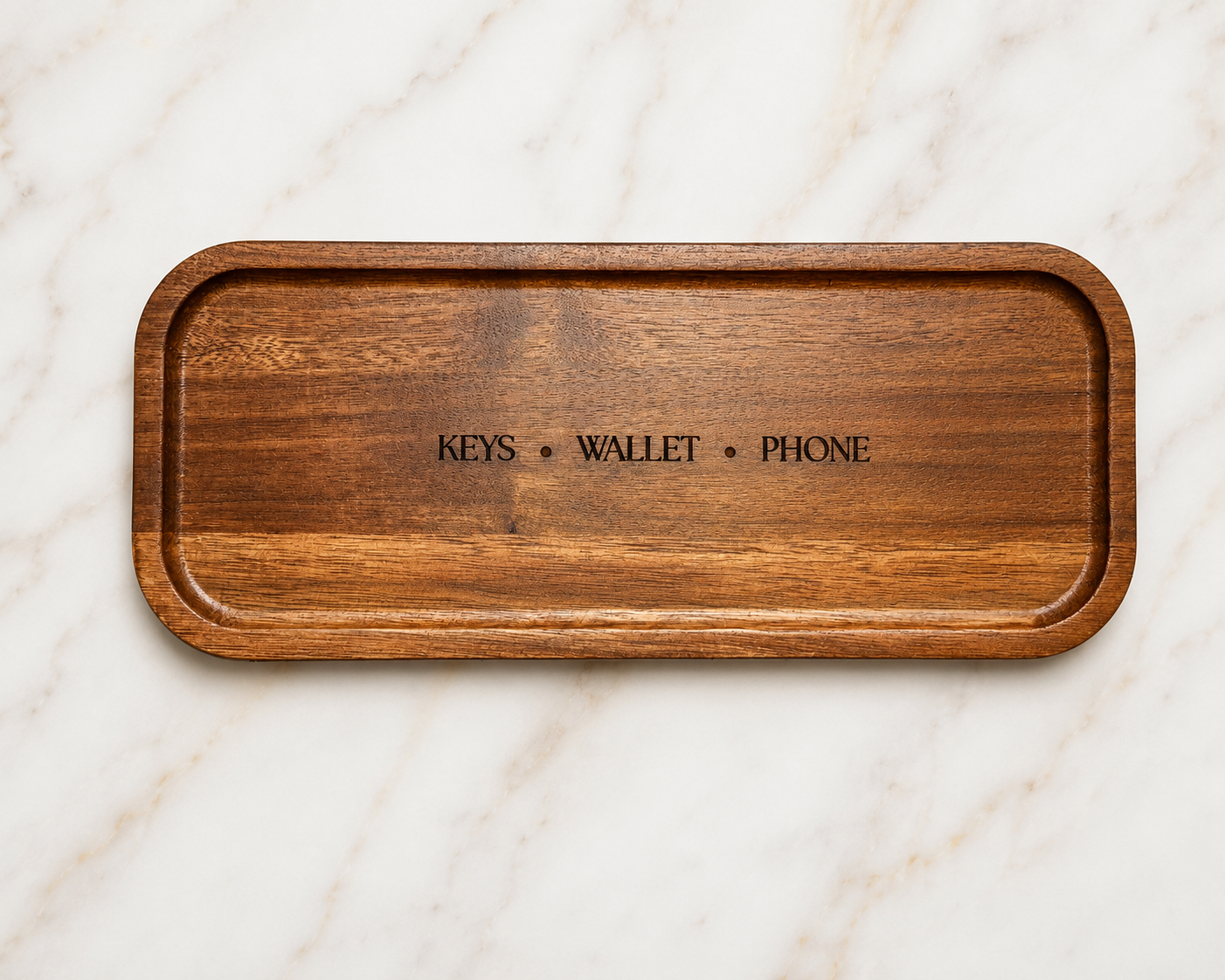 A rectangular wooden tray with rounded corners and embossed text reading 'KEYS • WALLET • PHONE' in the center, placed on a white marble surface.