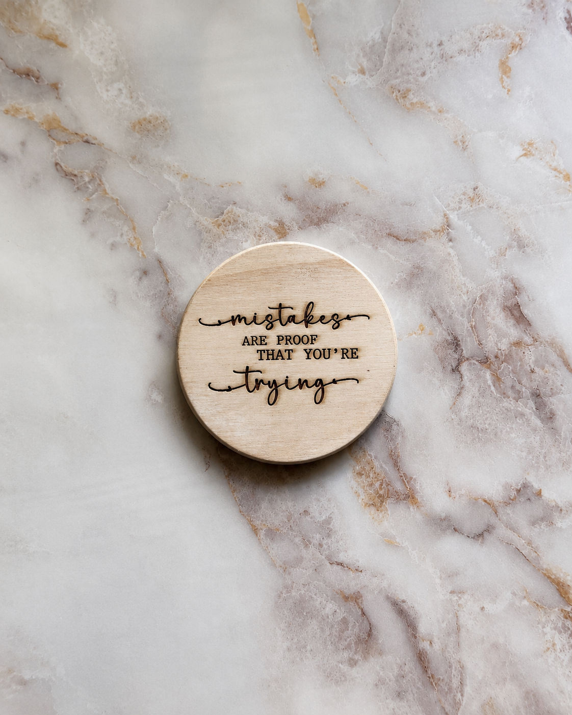 A round, light-colored wooden plaque with engraved black text reading 'mistakes ARE PROOF THAT YOU'RE trying', displayed on a white and beige marble surface.